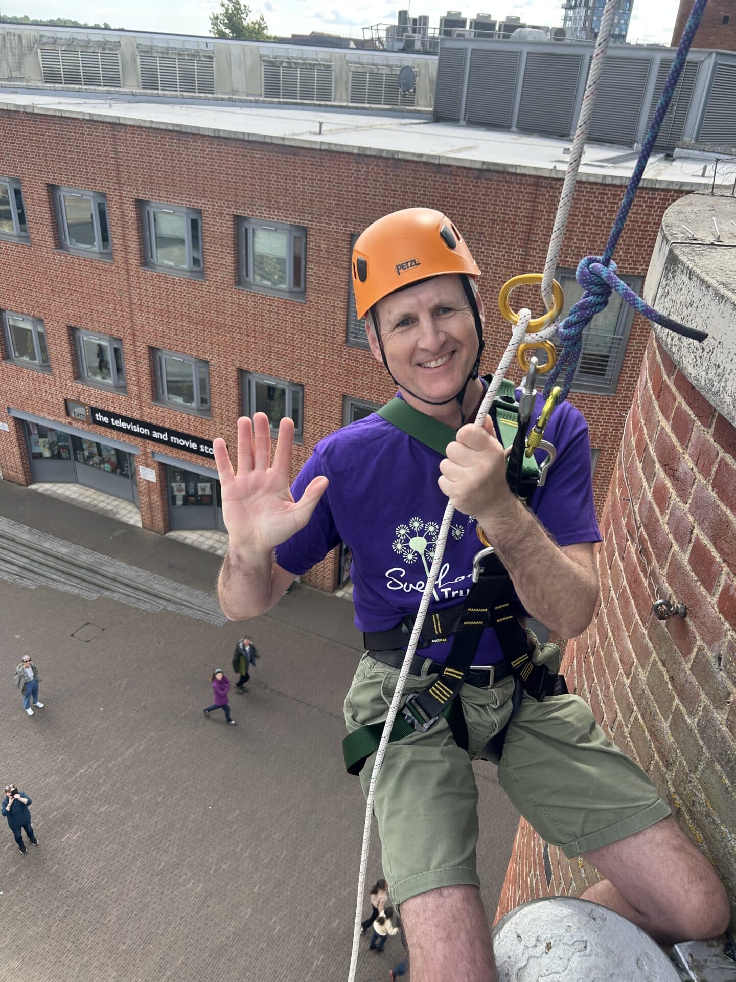 A man in an orange hard hat is attached to a building with ropes as he prepare to abseil down the Forum building in Norwich, wearing a Sue Lambert Trust purple t-shirt