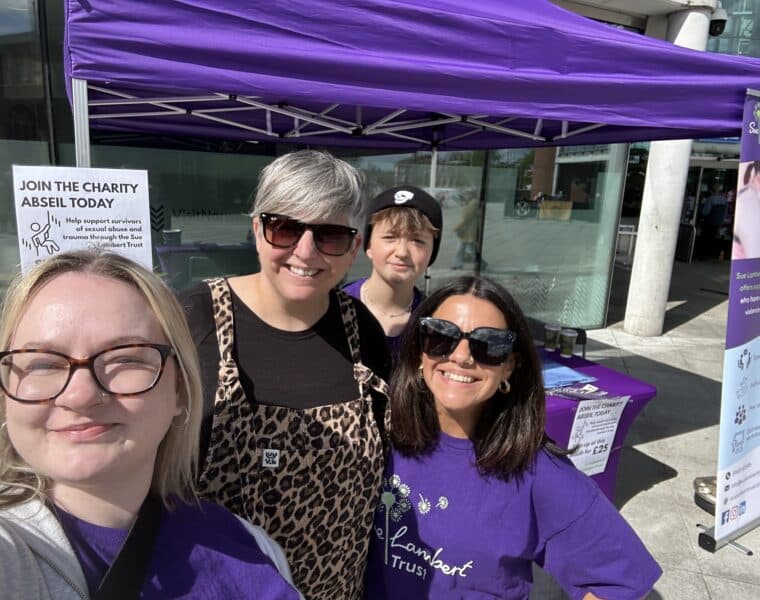 Four Sue Lambert Trust team members at a fundraising event standing together in branded purple t-shirts and standing in front of a purple gazebo, smiling