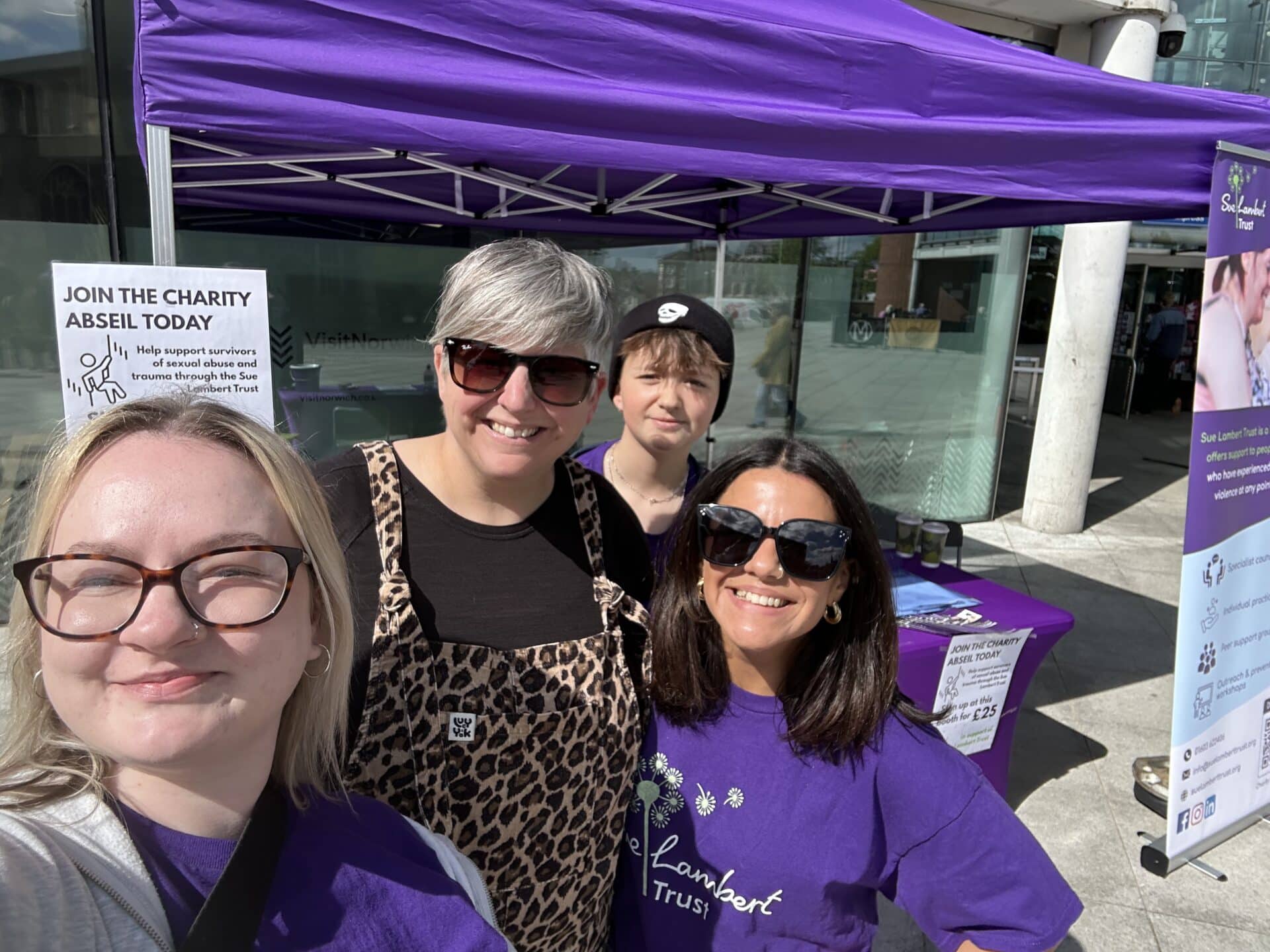 Four Sue Lambert Trust team members at a fundraising event standing together in branded purple t-shirts and standing in front of a purple gazebo, smiling
