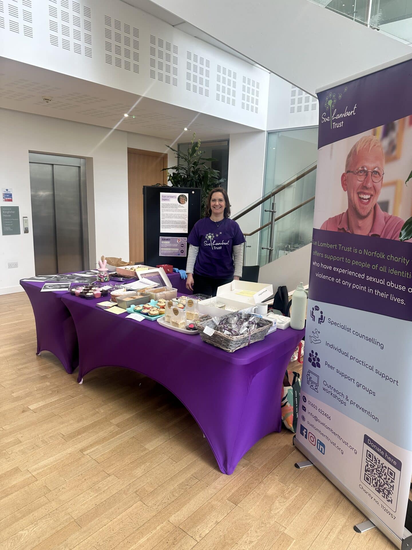 A bake sale stall in aid of Sue Lambert Trust showing homemade cakes on a table and a banner next to the table, explaining more about the work that the charity does to support survivors