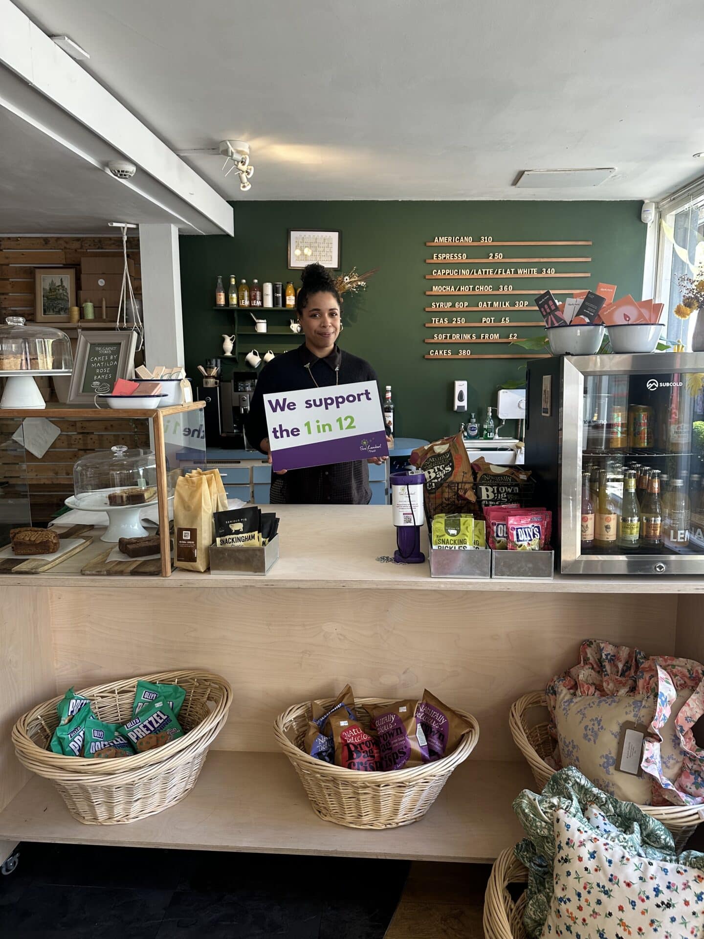 A team member at Tin Garnet Stores in Norwich is standing behind the serving counter holding a sign to show support for the Sue Lambert Trust